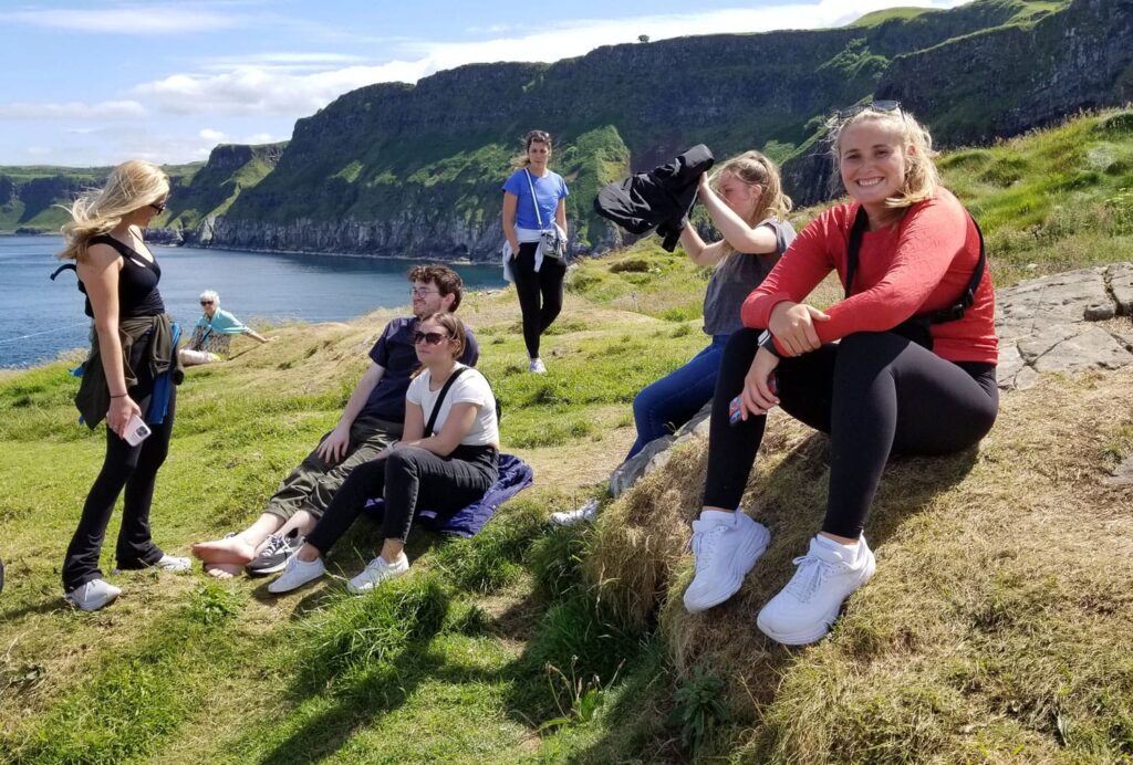 Students sit and stand on a grassy shore in Ireland on a student trip.