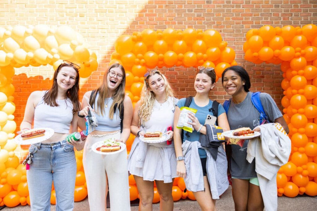 Five students hold plates of food while standing in front of orange balloons spelling out CCI.